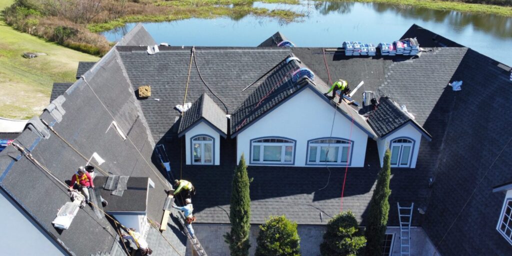 Roofing crew installing a large dark roof with dormers; shingle bundles are staged on the roof