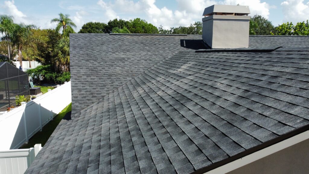 Gray architectural shingle roof with hip lines and a chimney under a partly cloudy sky