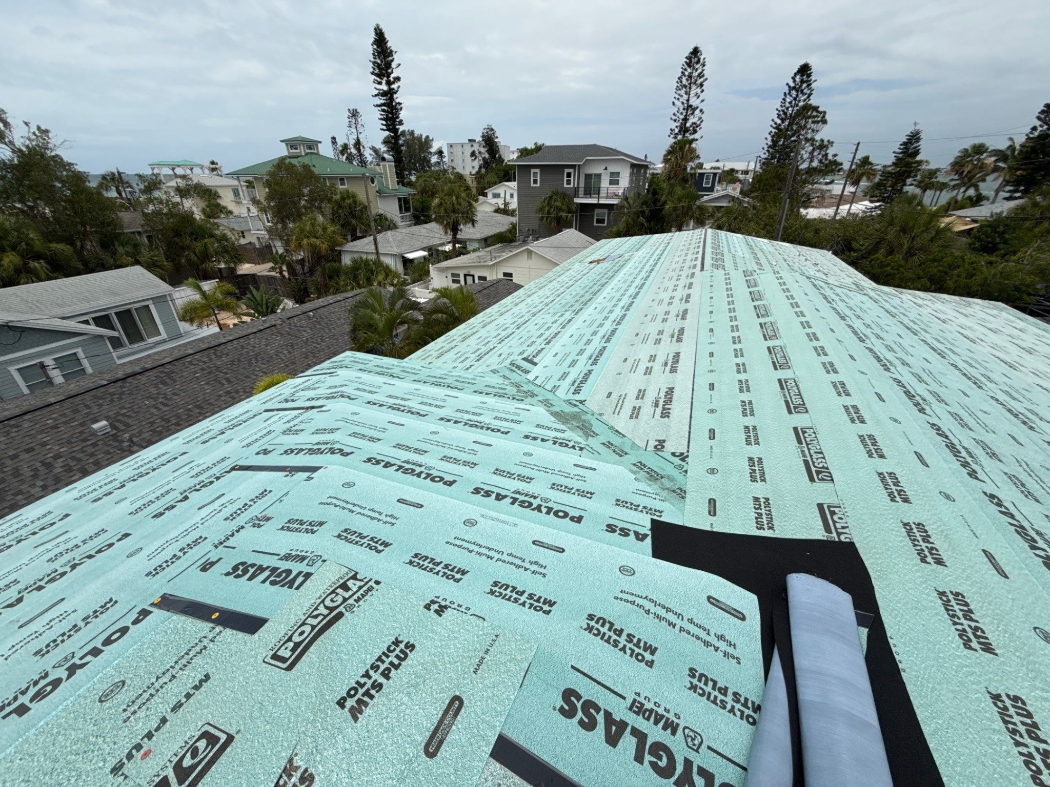 Overlapping synthetic underlayment sheets fastened across a roof deck with neighborhood rooftops in the background